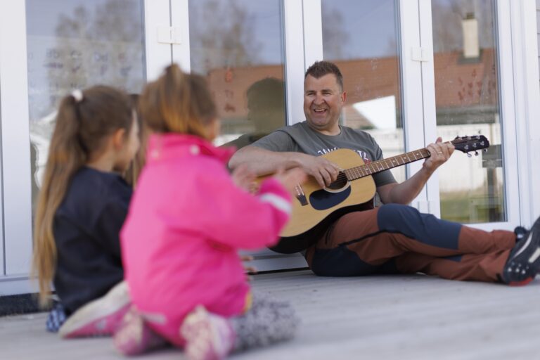 bildet viser mann sittende med gitar med to barn ved siden av seg